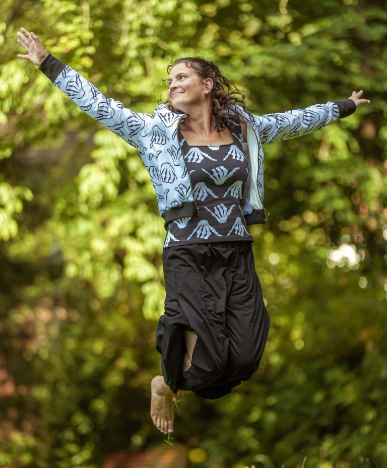 Frau in buntem Outfit springt fröhlich vor grünem Hintergrund.