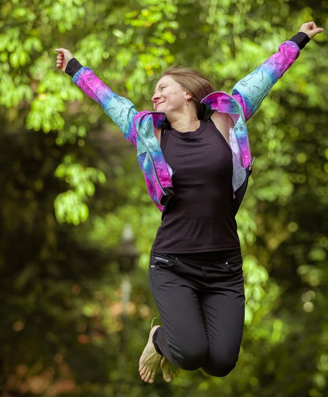 Frau in buntem Outfit springt fröhlich vor grünem Hintergrund.